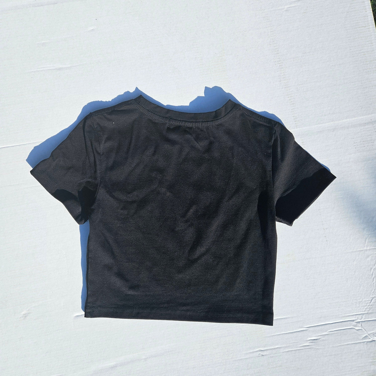 A black short-sleeve crop top shown from the back, laid flat on a white textured surface with a strong blue shadow.