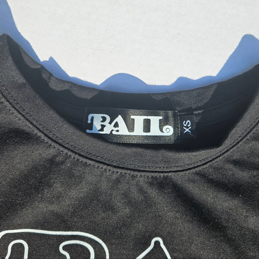 Close-up of a black t-shirt collar showing the 'BAIL' brand label and size 'XS' on a light grey background with defined shadows.