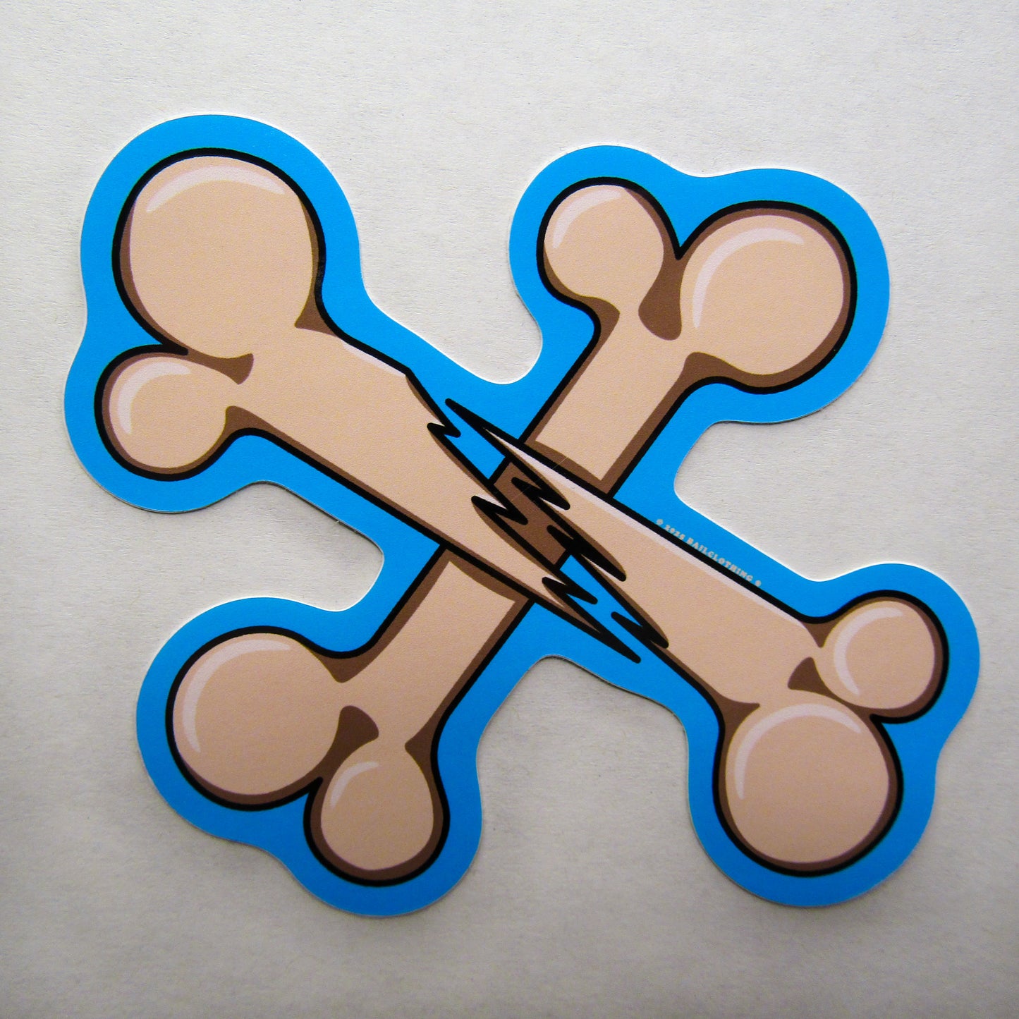 A single sticker of two crossed cartoon bones with a jagged break in the centre, named "BROKEN BONES". It is outlined in bright blue and stands against a white background, conveying a playful tone.