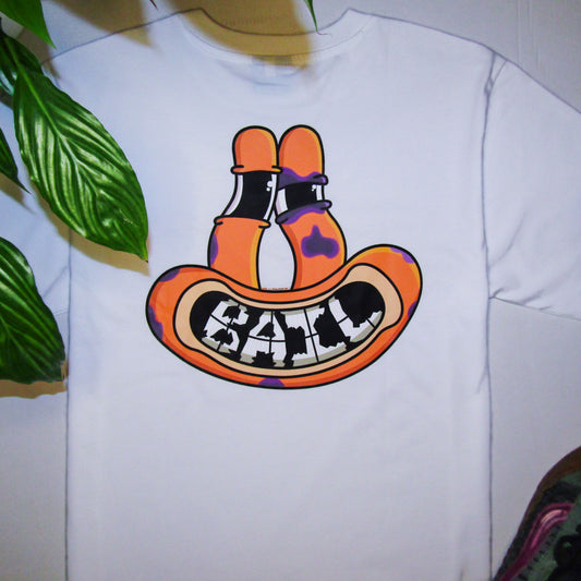 Lifestyle photograph featuring a white graphic T-shirt. Printed on the apparel is a colourful graphic design of an orange frog-like face with tall eyes. The face is smiling, revealing "BAIL" text compiled of shattered teeth, and has several purple bruises, one of which forms a black eye. The apparel is flat-laid on a white background framed by green foliage and a pair of green and purple Vans Knu Skool skating shoes.