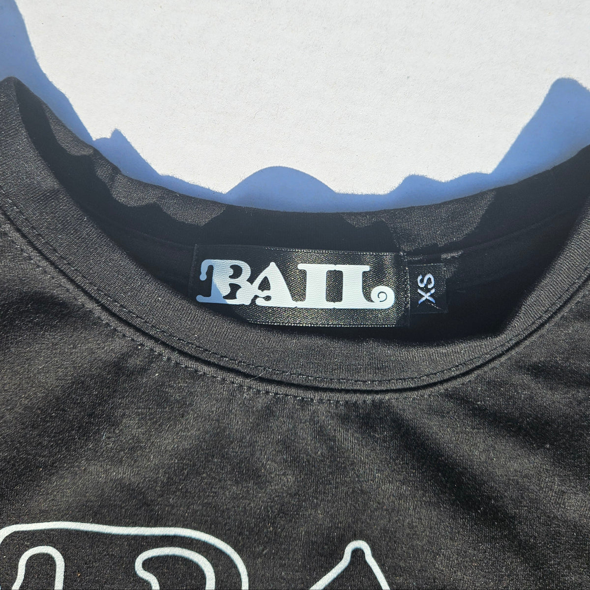 Close-up of a black t-shirt collar showing the 'BAIL' brand label and size 'XS' on a light grey background with defined shadows.