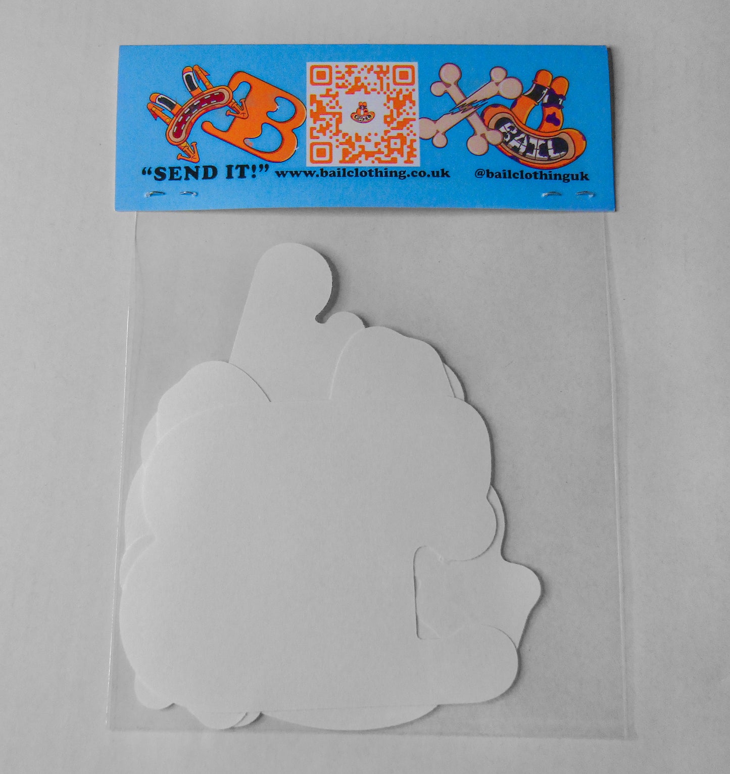 The back of a clear sticker pack bag is stapled closed with BAIL clothing's QR code, graphic designs of an orange frog and a website link. "SEND IT" is written in bold black lettering. The bag contains various white paper shapes showing the back of each sticker.