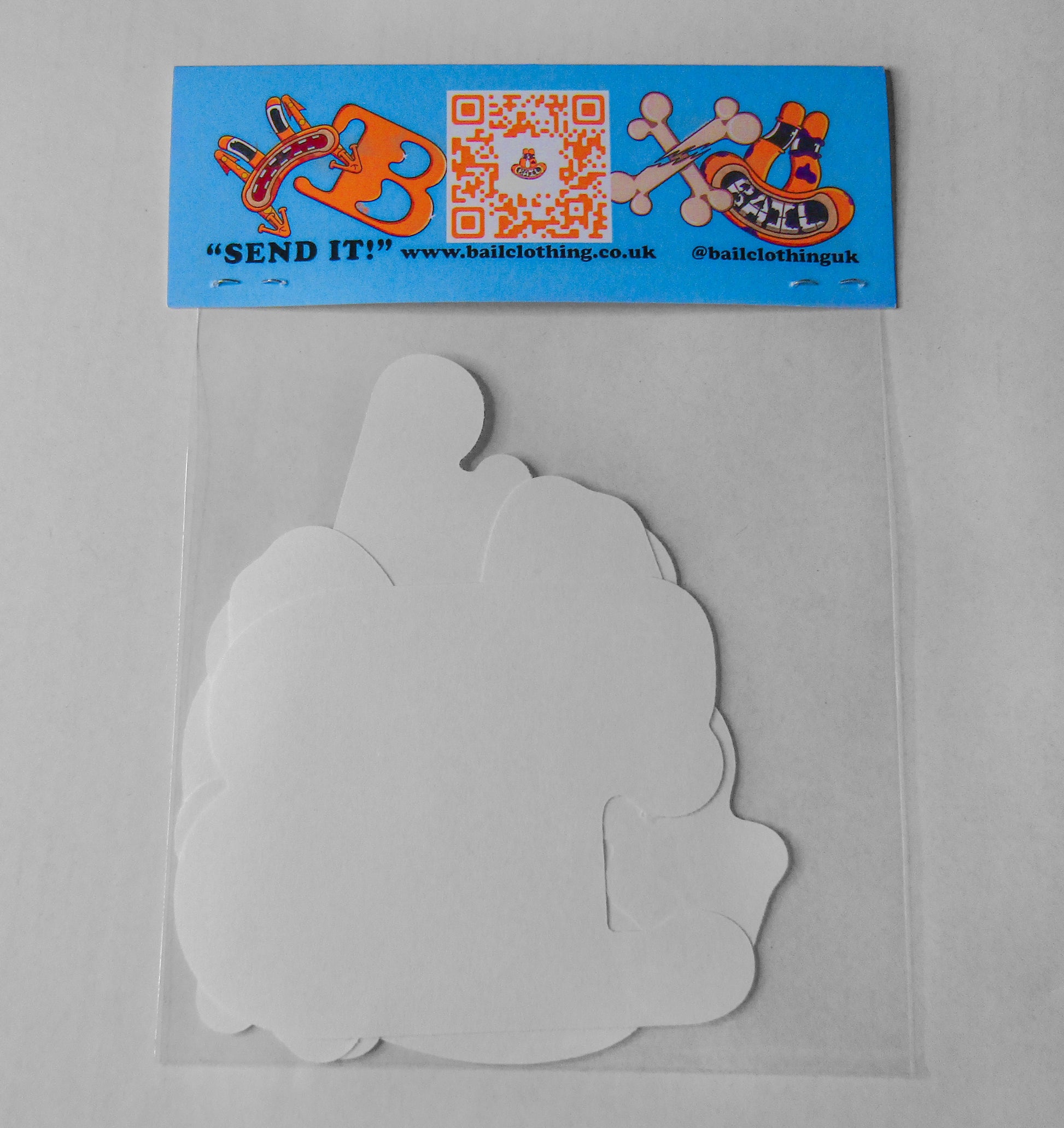 The back of a clear sticker pack bag is stapled closed with BAIL clothing's QR code, graphic designs of an orange frog and a website link. "SEND IT" is written in bold black lettering. The bag contains various white paper shapes showing the back of each sticker.