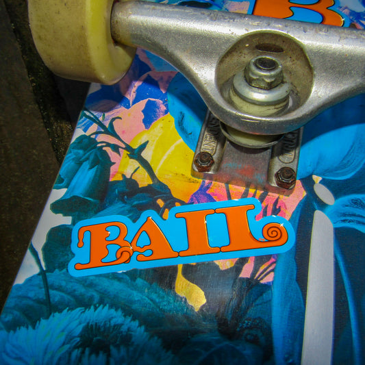 Close-up of a vibrant, primitive skateboard deck featuring multicoloured graphics and a sticker of the word and brand logo “BAIL” in bold, bubbly orange, stylised letters near a silver truck.
