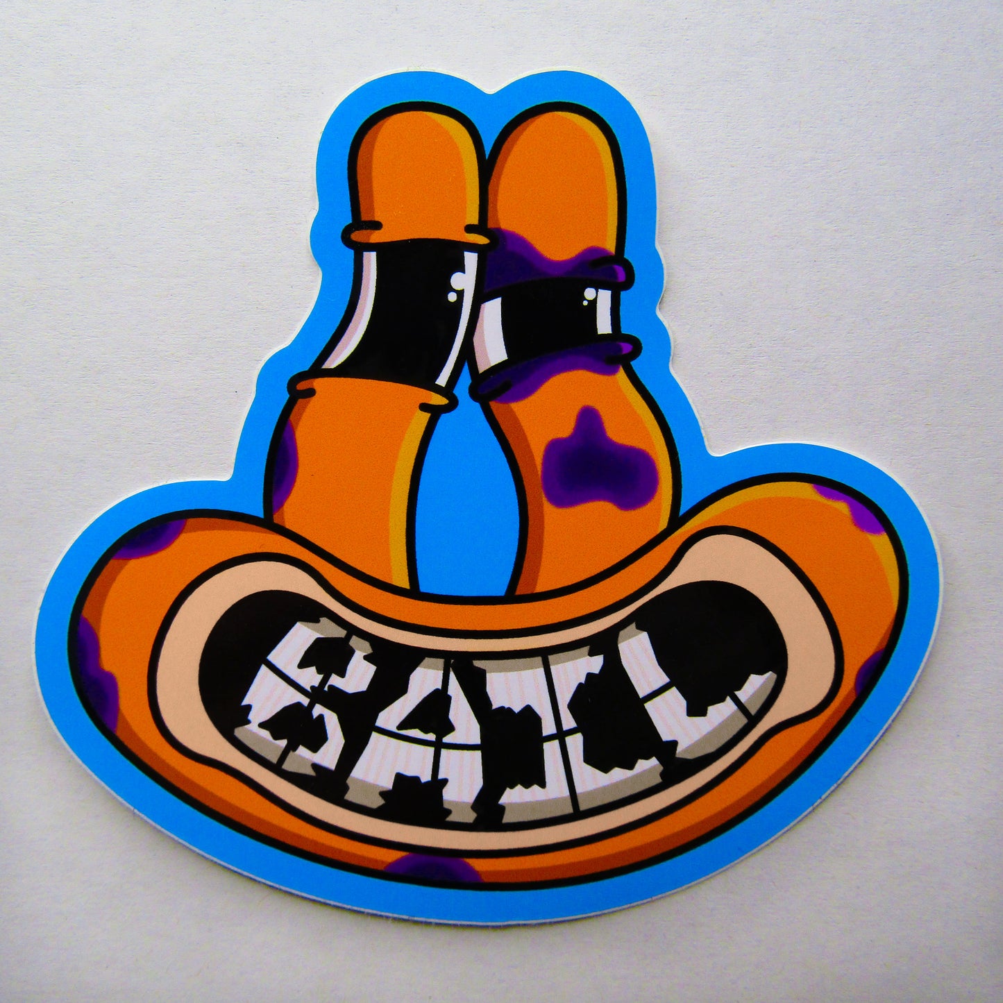 A singular sticker from a sticker pack of BAIL clothing. Cartoonish sticker showing an orange frog with long wobbly eyes, purple bruises scattering the face and making a black eye, and a wide mouth smiling to reveal the word "BAIL" on its teeth, set against a blue outline.