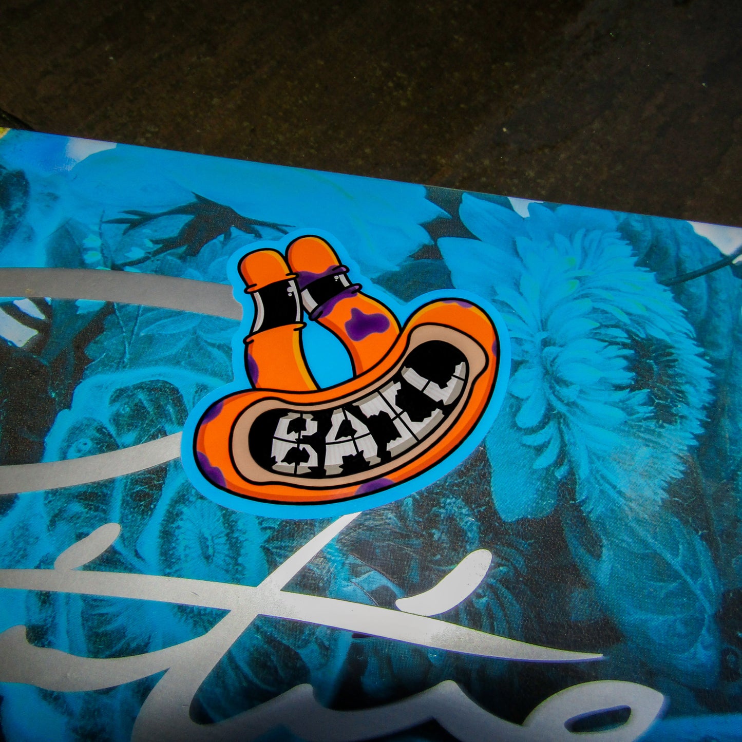 A zoomed in image of a primitive skateboard deck with bright yellow and blue hues. In the centre of the picture a colourful sticker showing an animated orange frog creature smiling with the word "BAIL" as its teeth stands out against a vibrant floral background, conveying a playful tone.