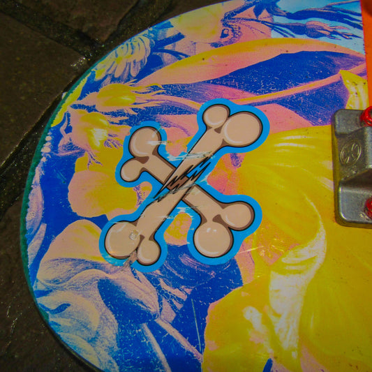 Close-up of the nose of a primitive skateboard deck which has vibrant yellow, blue, and pink hues, featuring a crossbones sticker in the centre. The white bones are broken in the middle and are surrounded by a light blue background. The mood is playful and artistic.