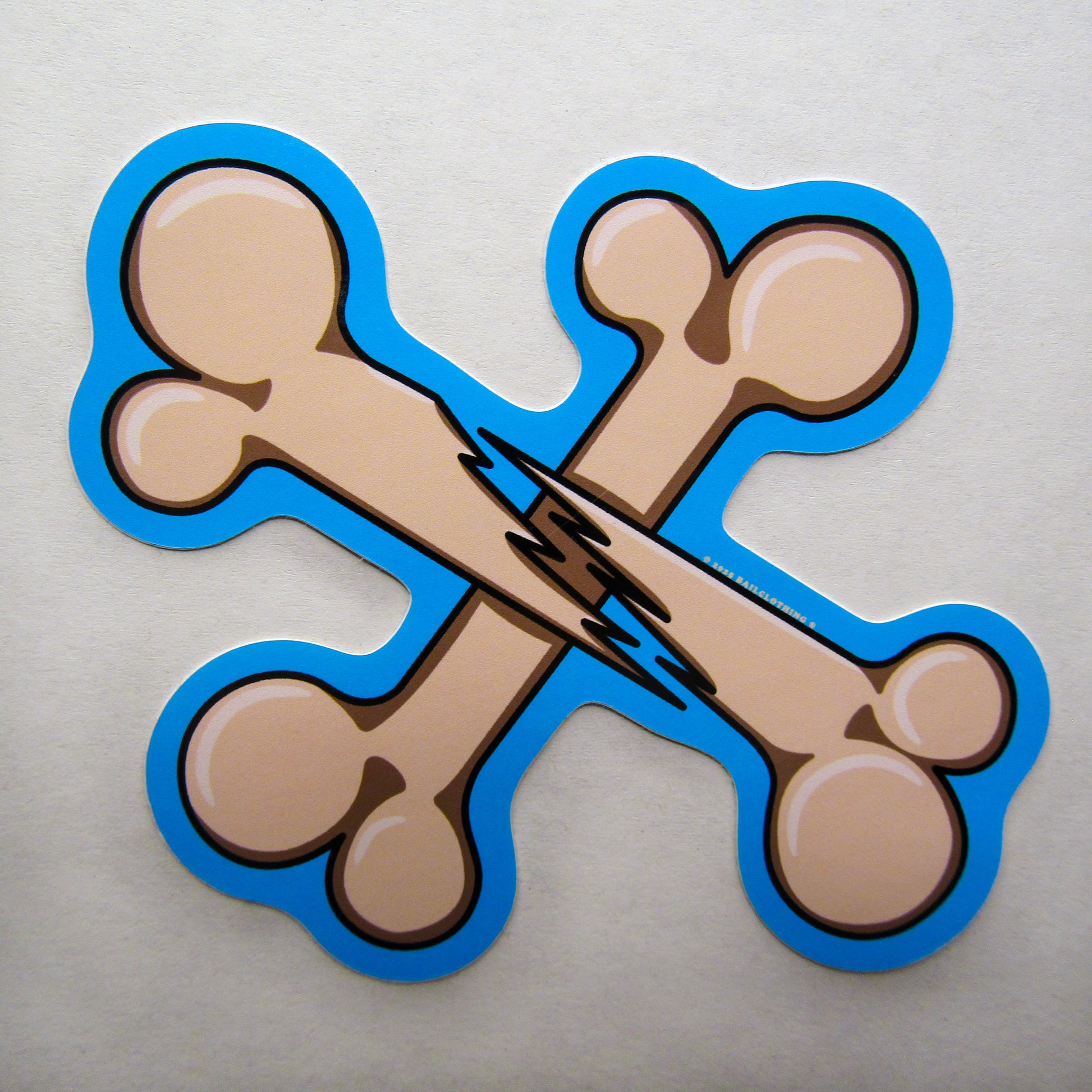 A single sticker of two crossed cartoon bones with a jagged break in the centre, named "BROKEN BONES". It is outlined in bright blue and stands against a white background, conveying a playful tone.
