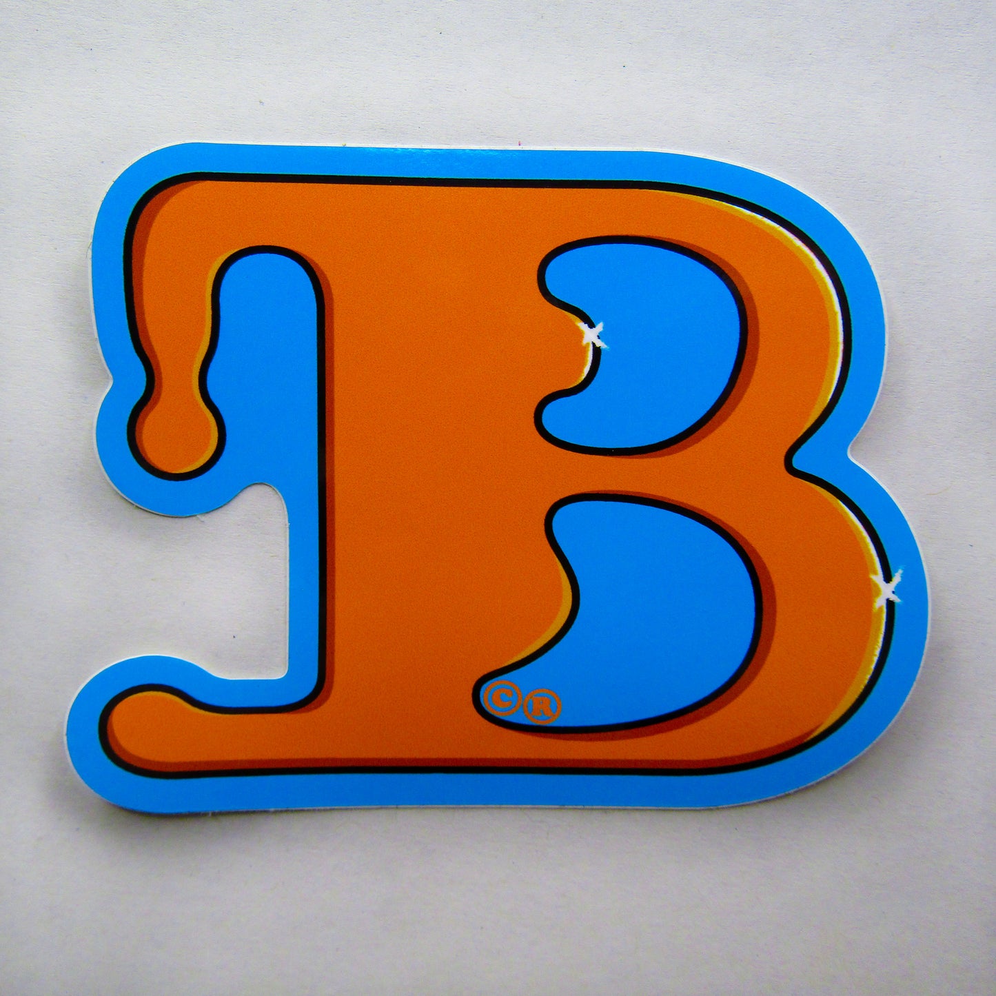 A singular sticker from the skateboarding brand 'BAIL'. Bold, stylised letter "B" in bright orange, outlined in light blue, resembling drippy paint. White strokes glisten from the curves of the font style. The design feels playful and vibrant.
