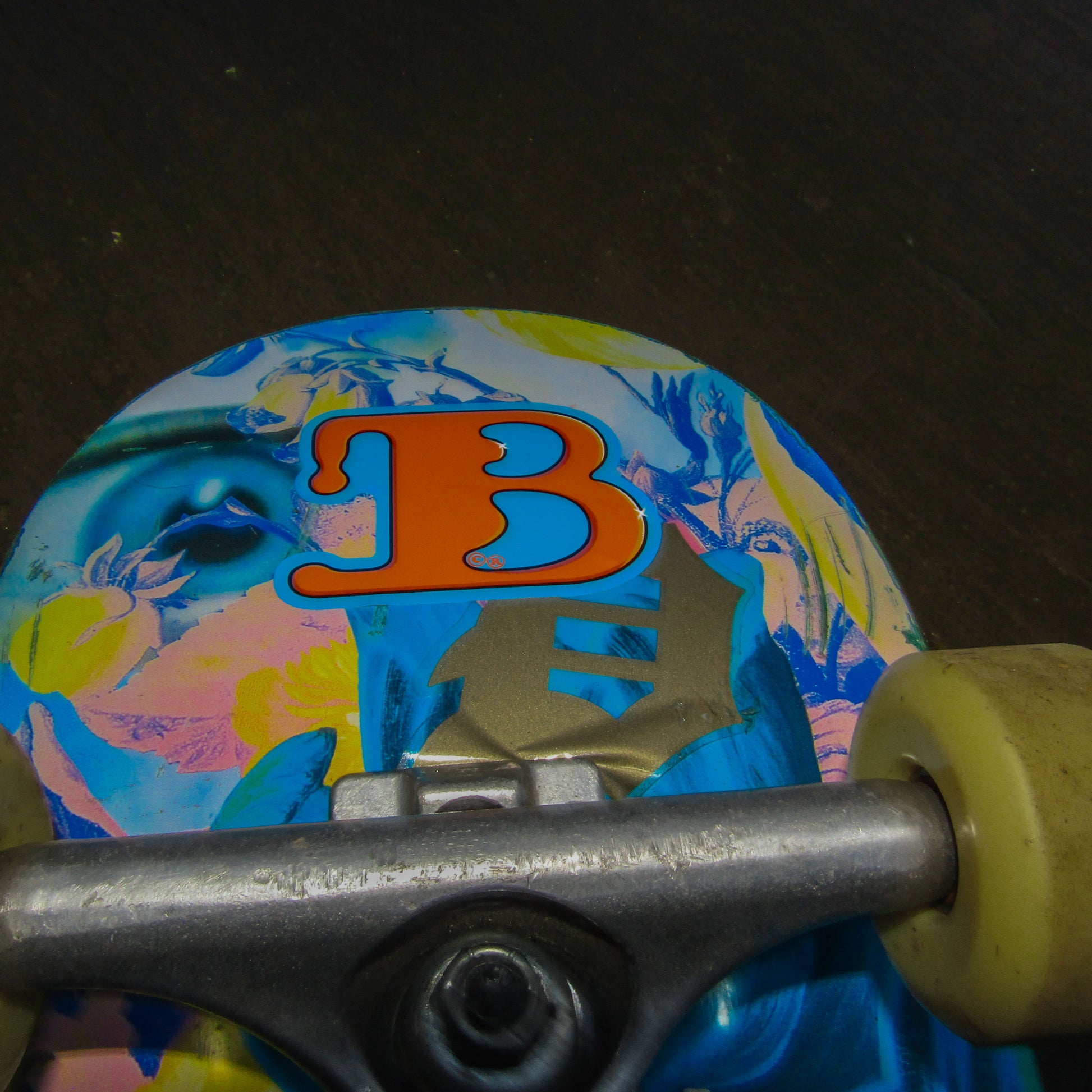 Close-up of a colourful primitive skate brand skateboard deck with a vibrant floral design featuring yellow and blue hues with an eye merged in. An orange "B" for the streetwear brand 'BAIL' clothing stands out prominently at the centre, showing that the board user has stuck on a bail sticker.