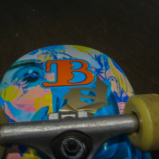 Close-up of a colourful primitive skate brand skateboard deck with a vibrant floral design featuring yellow and blue hues with an eye merged in. An orange "B" for the streetwear brand 'BAIL' clothing stands out prominently at the centre, showing that the board user has stuck on a bail sticker.