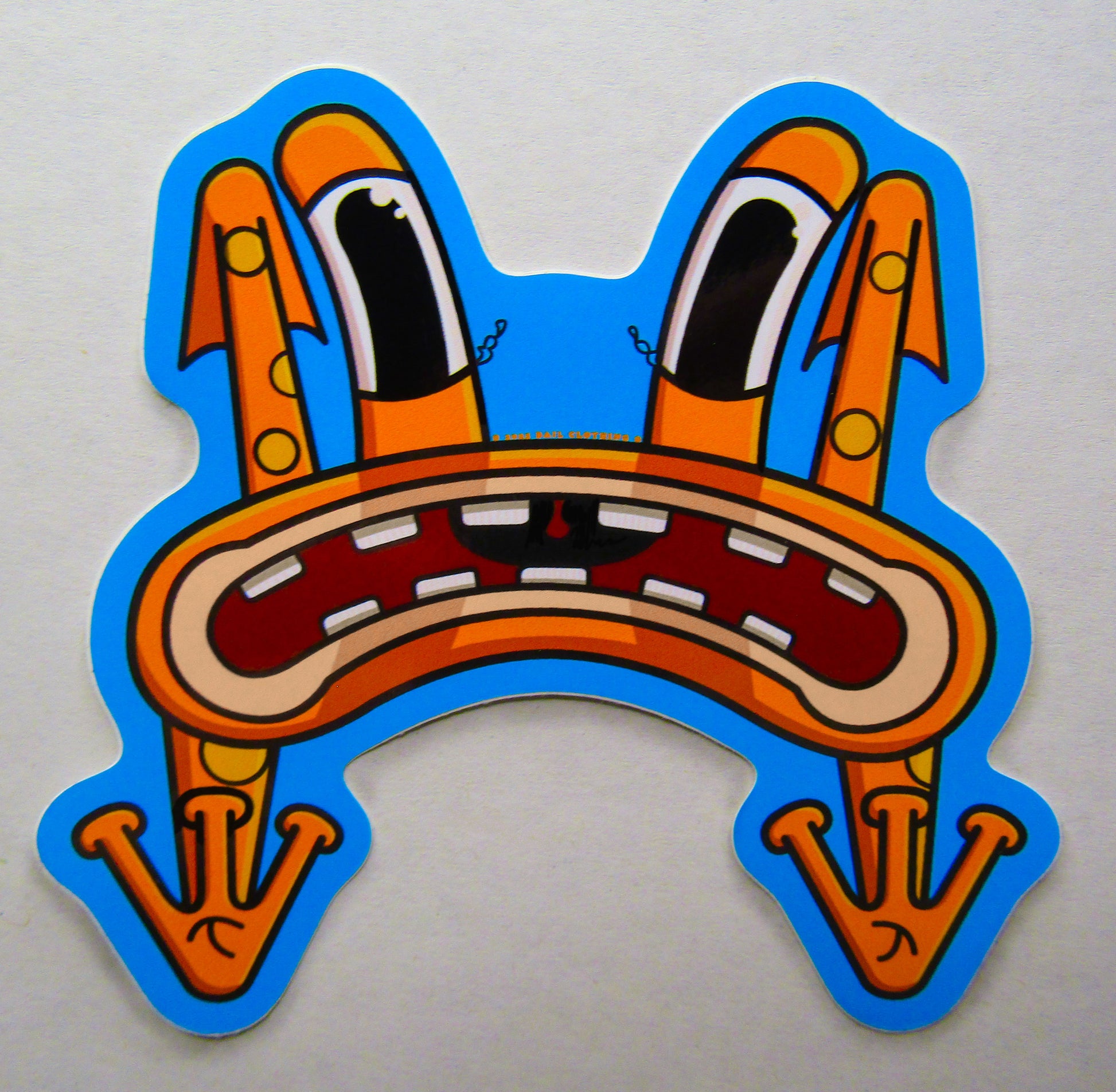 A close-up picture of a single sticker from BAIL clothing's sticker pack. Cartoonish, orange frog character with long eyes, an exaggerated frown, and whimsical design flying through the air as it gets thrown off of a skateboard in the skatepark. It conveys a humorous, quirky and relatable expression.