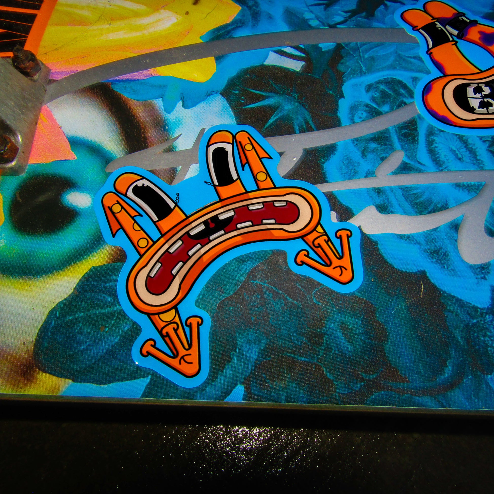 A picture of a primitive skate brand skateboard deck with a vibrant floral design showing strong yellow and blue colours. A cartoon-style sticker of an expressive, orange character frog with long eyes and a wide, frowning open mouth is shown flying to the ground from skating and bailing. Bright and lively tone.
