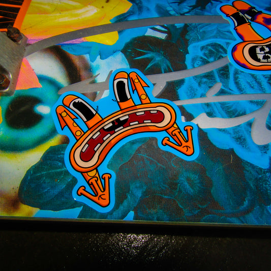 A picture of a primitive skate brand skateboard deck with a vibrant floral design showing strong yellow and blue colours. A cartoon-style sticker of an expressive, orange character frog with long eyes and a wide, frowning open mouth is shown flying to the ground from skating and bailing. Bright and lively tone.
