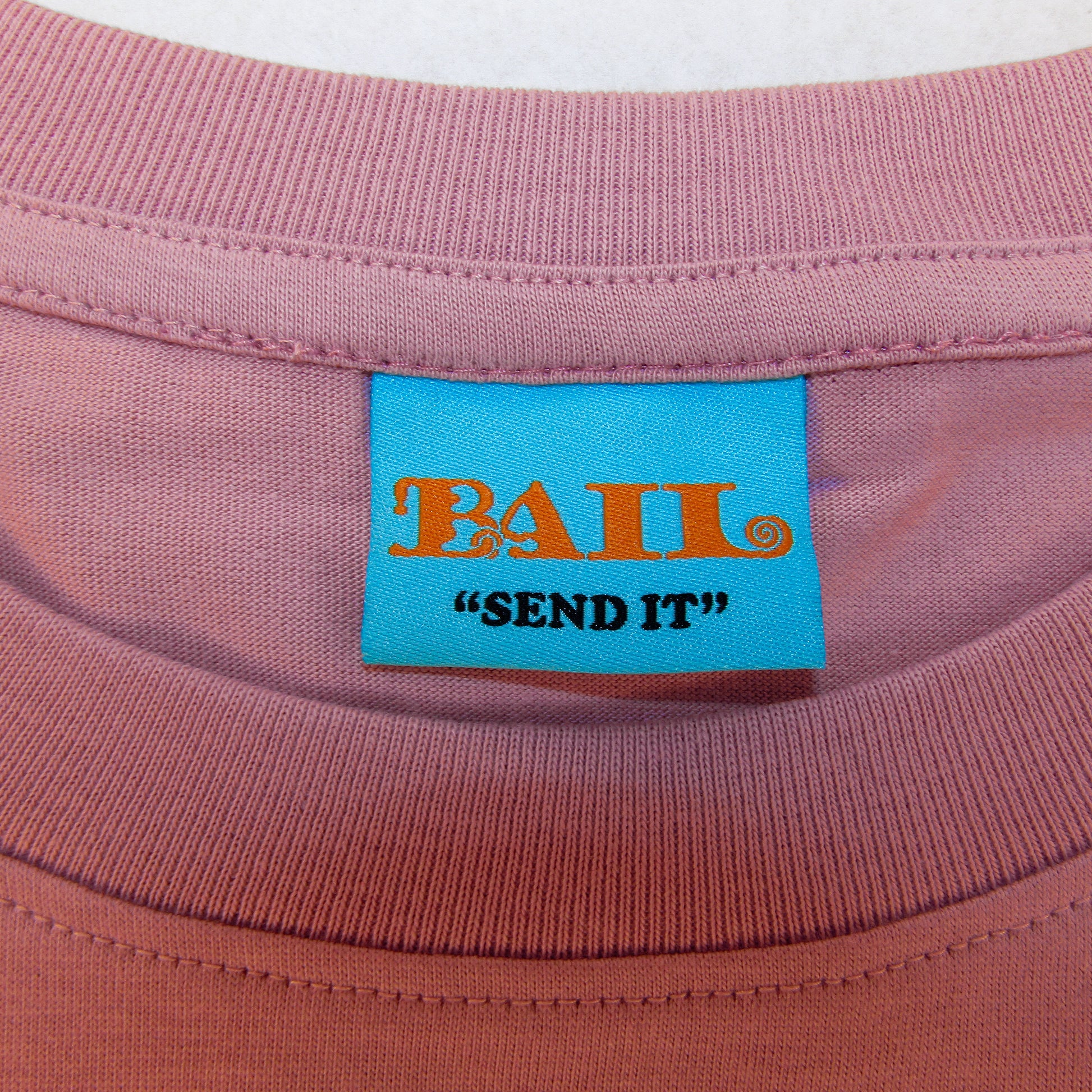 A close-up of a greyish-pink T-shirt collar with a bright blue label. The label features orange, ornate text reading "BAIL" and black text below saying the streetwear brand's motto "SEND IT."