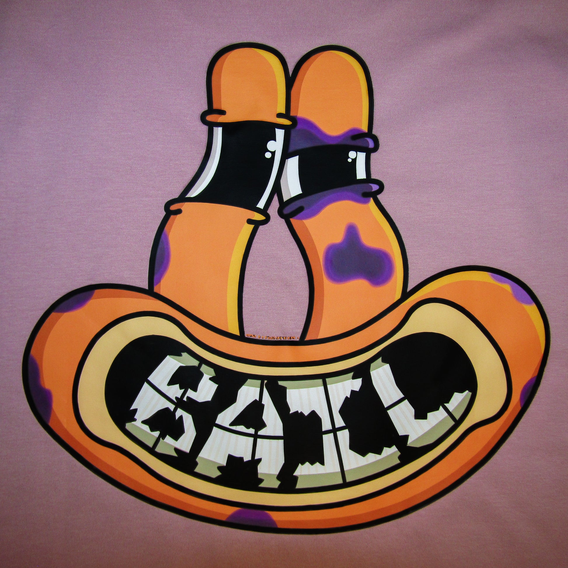 Colourful cartoon art on the back of a pink 100% cotton tee featuring an orange alien frog face with tall eyes and a wide, black-and-white grinning mouth with cracked teeth spelling the clothing brand's name,  "BAIL." The face has purple bruises and a black eye, indicating the frog fell while skateboarding, yet it has a happy, relaxed expression.
