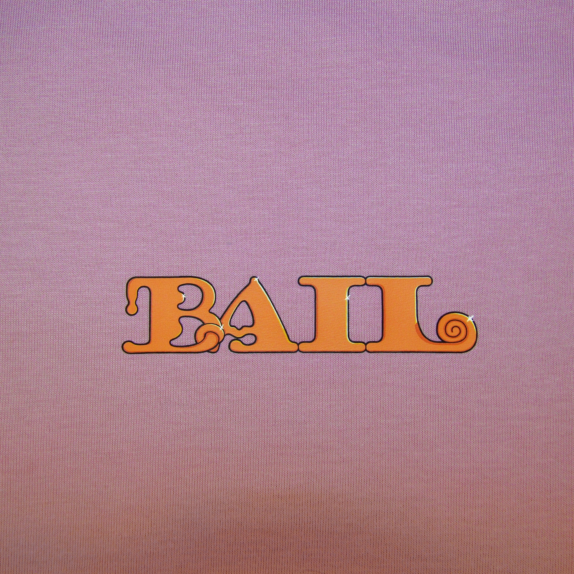 A close-up shot of the "BAIL" logo on the front left of the dusty pink tee. The text is centred in the image and shows the retro, orange font with swirls, creating a vintage, playful tone.