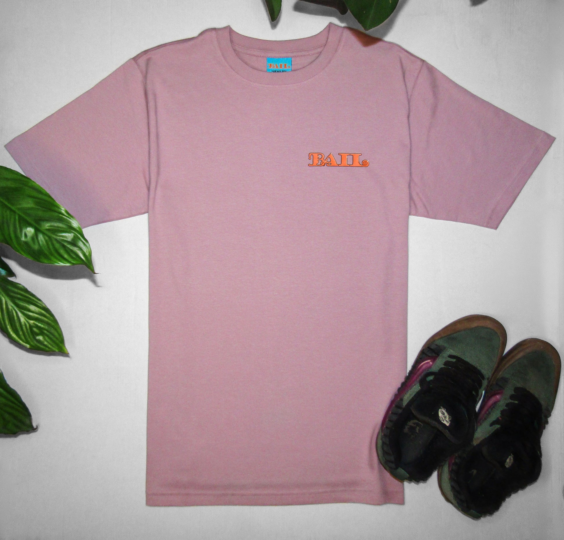 Lifestyle photography showing a dusty pink oversized T-shirt with orange "BAIL" text in a bold, bubbly font and a sky-blue neck tag. It is flat-laid on a white background, surrounded by green plant leaves and a pair of green and purple Vans Knu Skool skateboarding shoes. Casual and stylish.