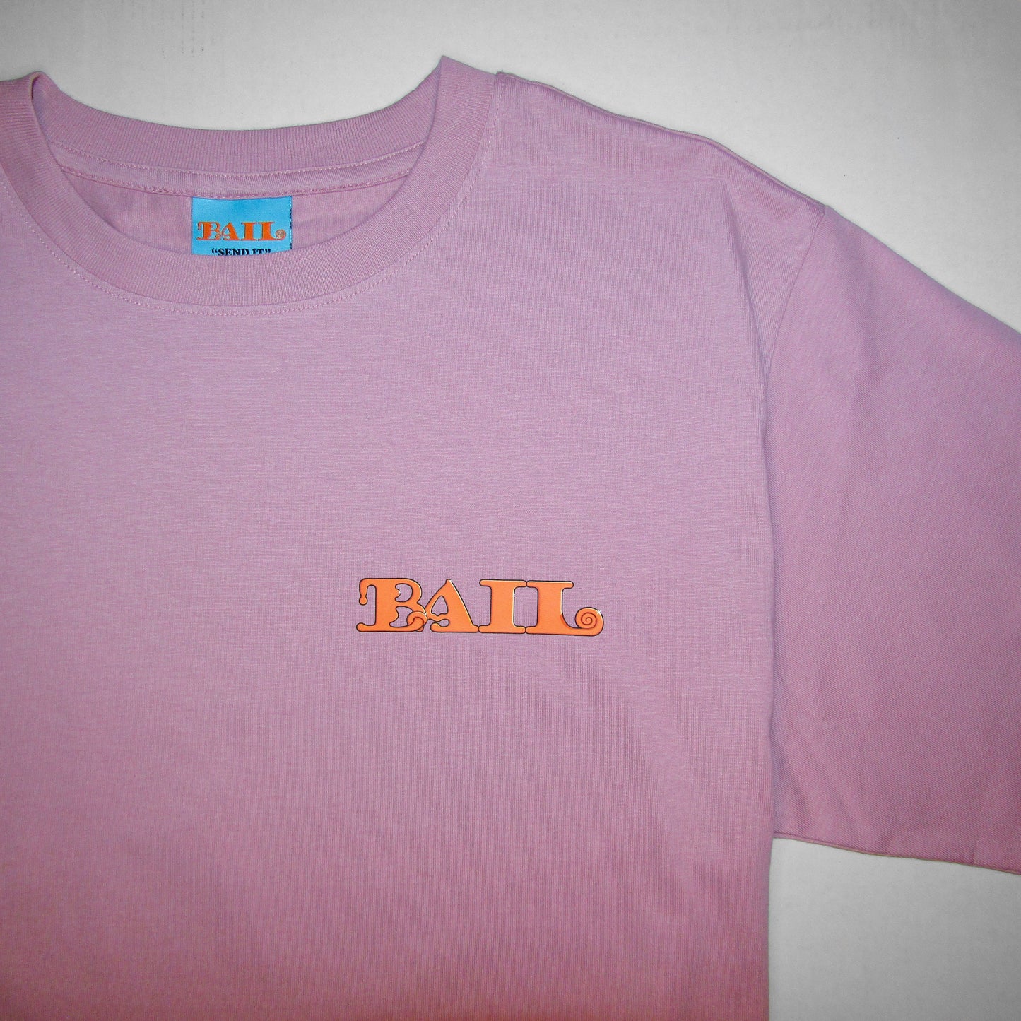 A close-up of a light pink T-shirt with a round neck, featuring UK clothing brand Bail's bold orange "BAIL" text logo on the left chest in a rounded font and a light blue woven neck tag with the same logo and Bail's brand motto "Send It" in black. The apparel evokes a casual, trendy vibe, accentuated by simple colour contrasts.
