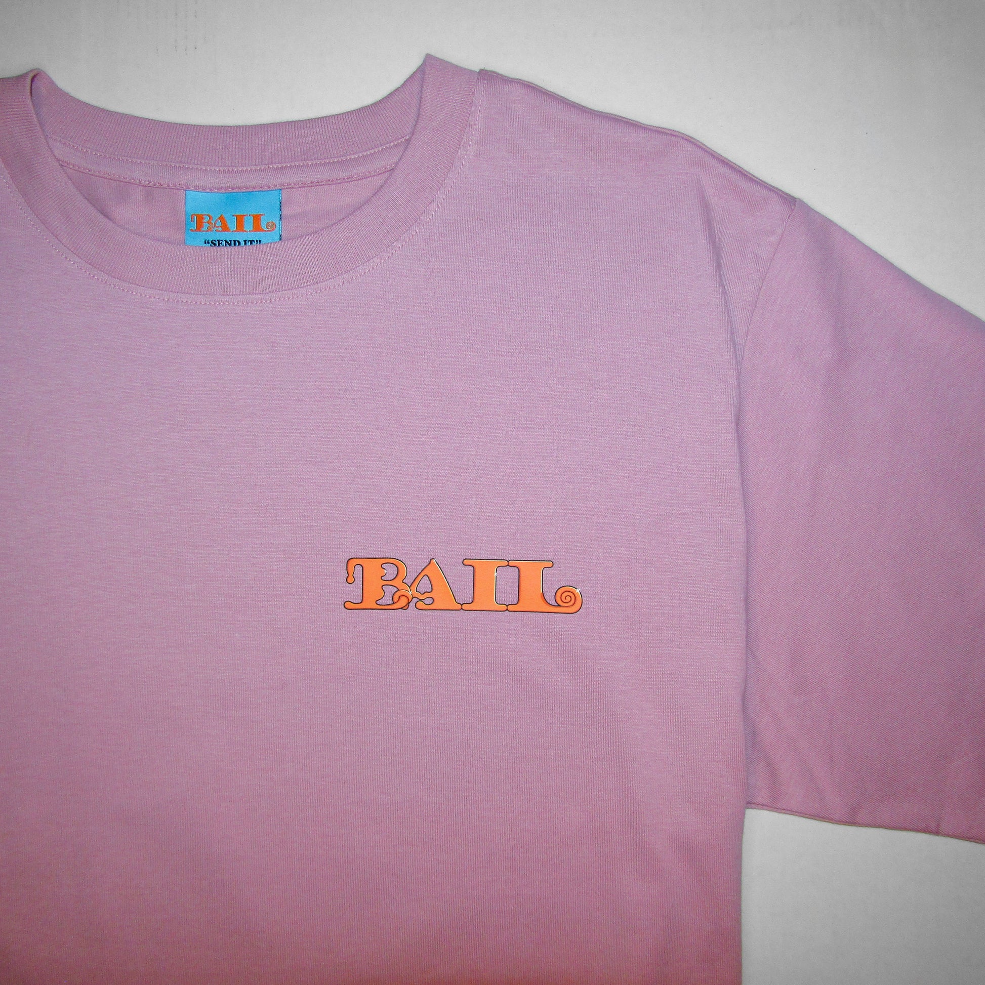 A close-up of a light pink T-shirt with a round neck, featuring UK clothing brand Bail's bold orange "BAIL" text logo on the left chest in a rounded font and a light blue woven neck tag with the same logo and Bail's brand motto "Send It" in black. The apparel evokes a casual, trendy vibe, accentuated by simple colour contrasts.
