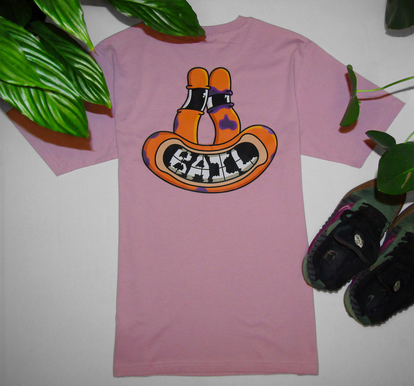 Lifestyle photography showing a light pink graphic T-shirt featuring a colourful cartoon face design of an orange frog-like character with tall eyes. The face is smiling, revealing "BAIL" text compiled of broken teeth, and has several purple bruises, one of which forms a black eye. The apparel is flat-laid on a white background framed by green foliage and a pair of green and purple Vans Knu Skool skating shoes.