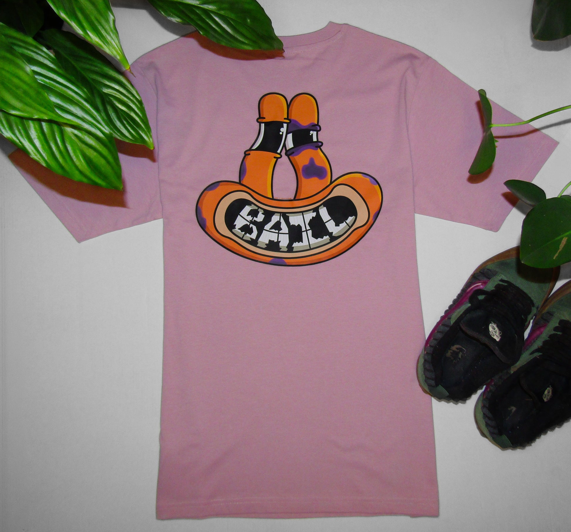 Lifestyle photography showing a light pink graphic T-shirt featuring a colourful cartoon face design of an orange frog-like character with tall eyes. The face is smiling, revealing "BAIL" text compiled of broken teeth, and has several purple bruises, one of which forms a black eye. The apparel is flat-laid on a white background framed by green foliage and a pair of green and purple Vans Knu Skool skating shoes.