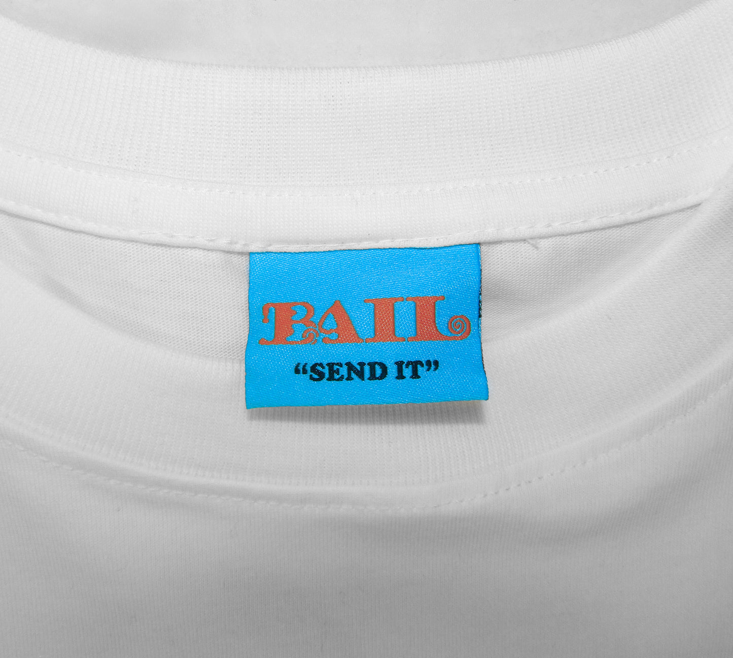 A close-up shot shows a bright blue clothing label on a white t-shirt with a crew neck. It displays the brand name "BAIL" in orange bubbly font with a swirl in the L, along with the skate brand's motto "SEND IT" in black, conveying a casual, playful tone.
