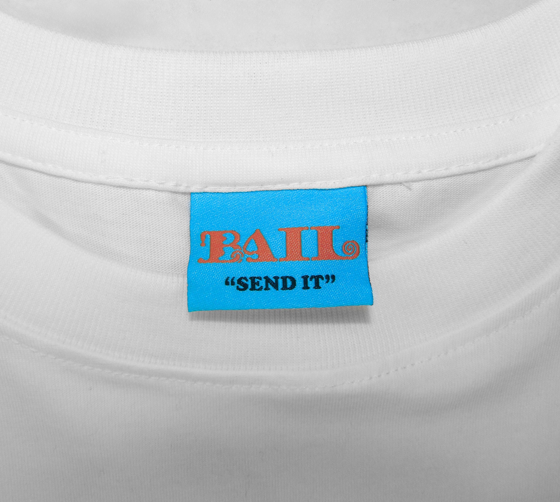 A close-up shot shows a bright blue clothing label on a white t-shirt with a crew neck. It displays the brand name "BAIL" in orange bubbly font with a swirl in the L, along with the skate brand's motto "SEND IT" in black, conveying a casual, playful tone.
