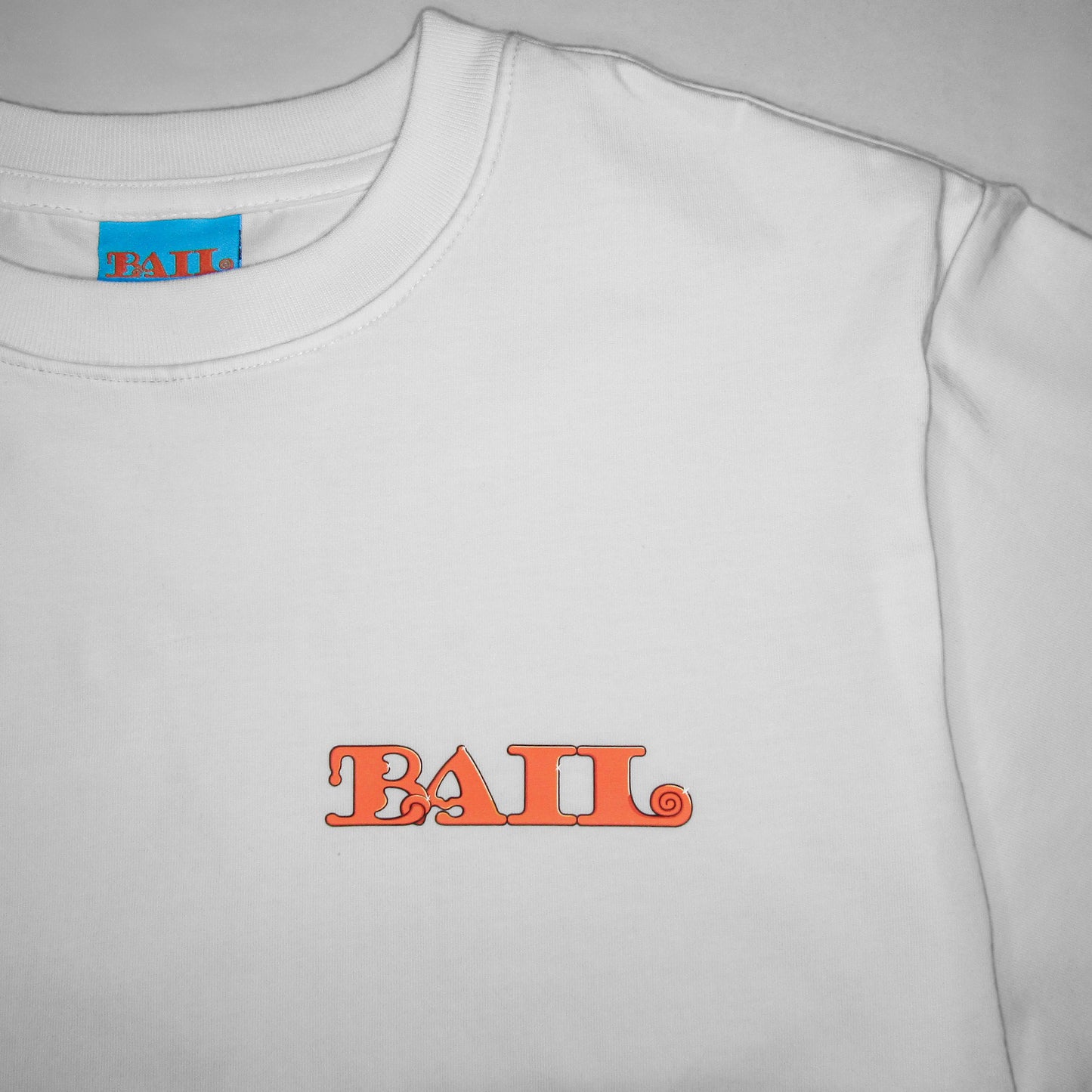 Close-up photography of a white T-shirt with a round neck and the word "BAIL" printed in bold, orange retro-style letters on the front. The tag on the apparel is bright blue with matching orange text.