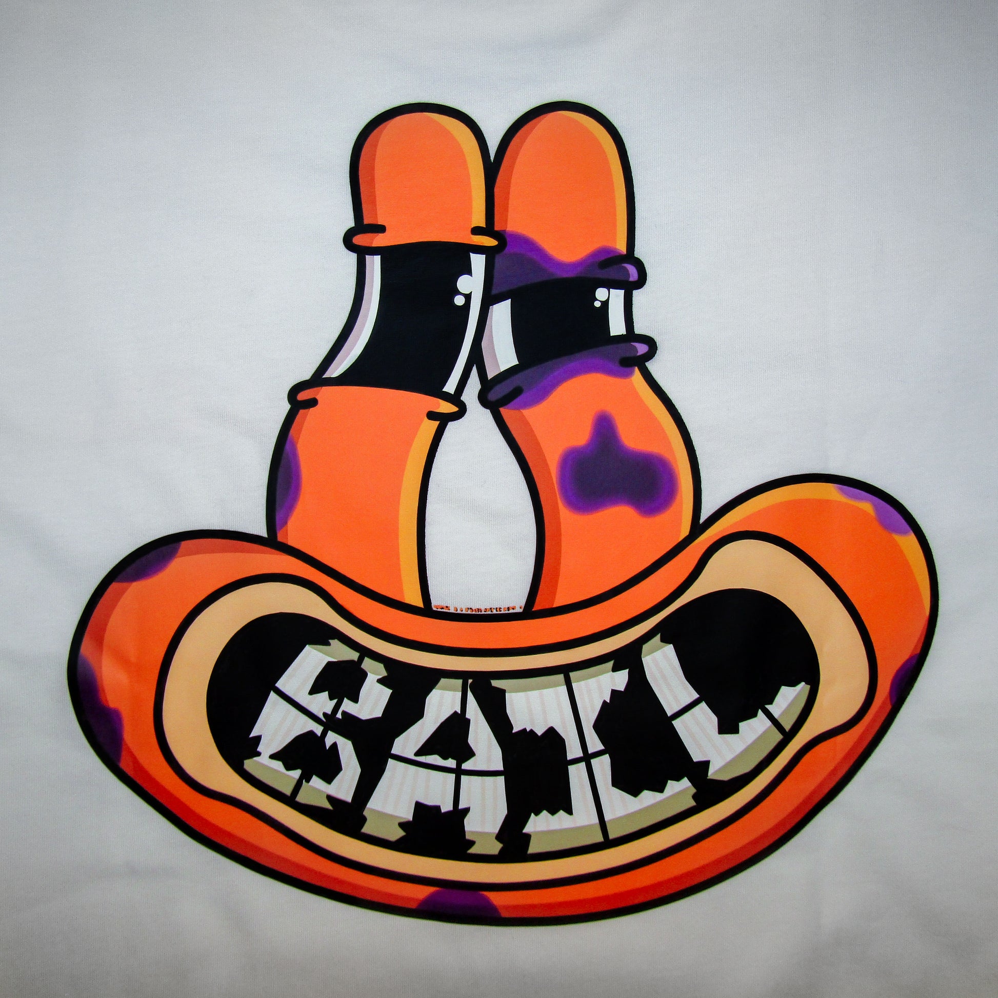 Close up of a colourful cartoon direct to film print on the back of a white t-shirt featuring an orange alien frog face with tall eyes and a wide, black-and-white grinning mouth with cracked teeth spelling the brand name, "BAIL." The face has purple bruises and a black eye, indicating the frog fell while skating.