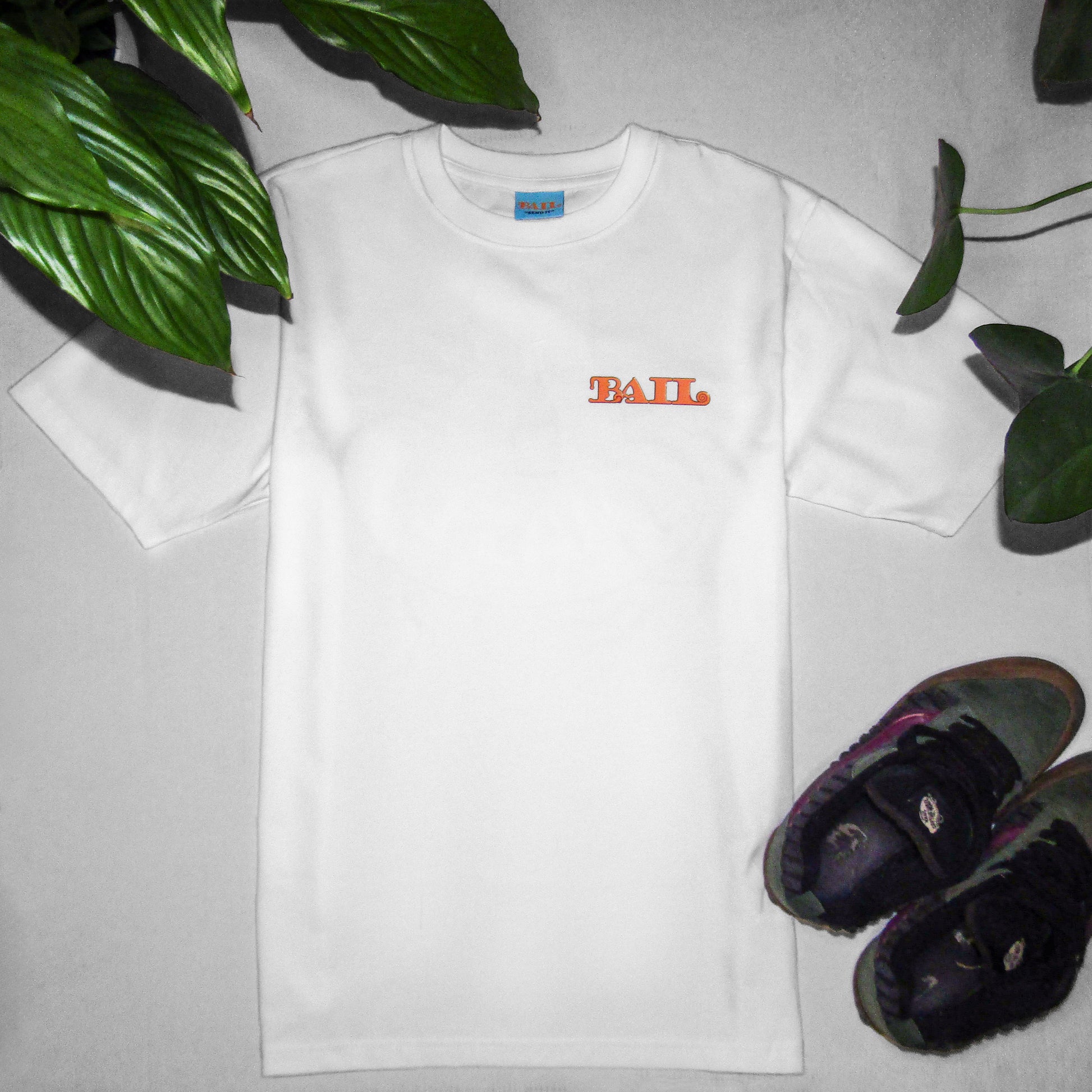An oversized white streetwear style T-shirt with a crew neck shows "BAIL" in orange curvy text on the left chest, laid flat on a white background. It is surrounded by vibrant green plant leaves and a pair of colourful green and purple Vans Knu Skool skateboarding shoes. The tone is casual and fresh.