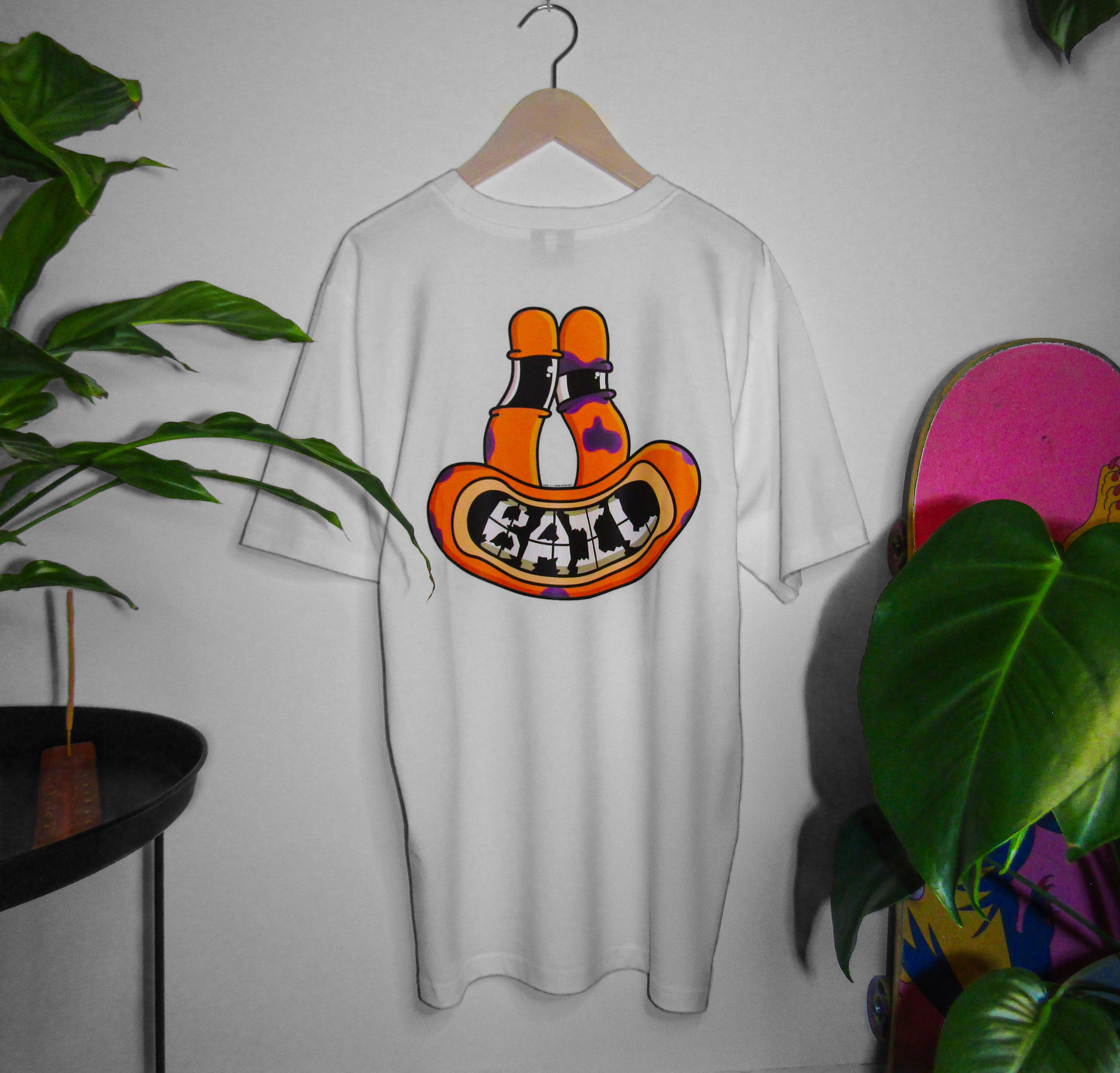 Lifestyle photography featuring a 100% cotton white streetwear t-shirt with "BAIL" in a bold,  bubbly orange font. The apparel is hung on a white wall by a wooden clothes hanger and is framed by green plants, a Japanese peace lily and a monstera; a pink skateboard is partially visible, along with an incense burner on a black table. The vibe is calm and cosy.
