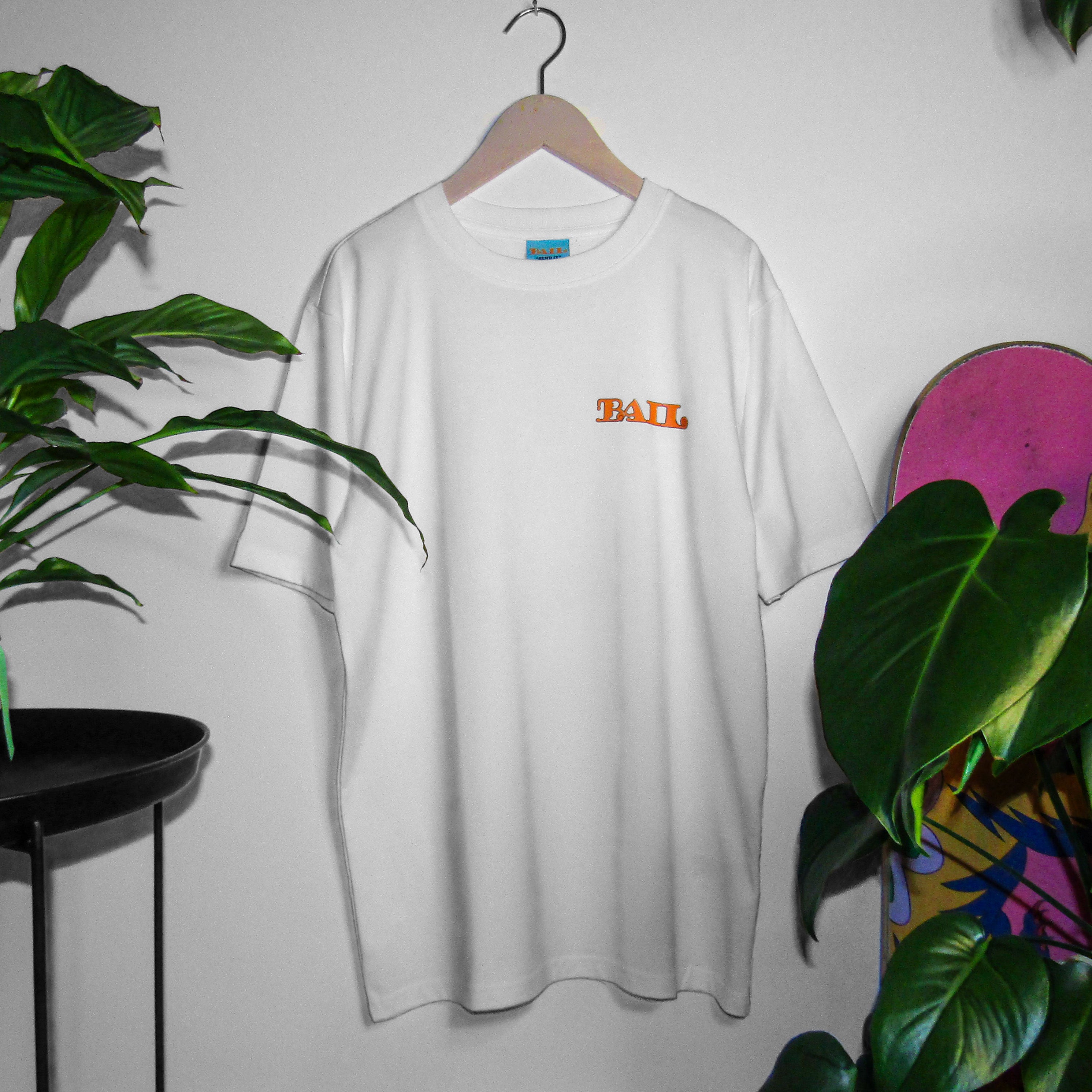 Lifestyle photography featuring a 100% cotton white streetwear t-shirt with "BAIL" in a bold,  bubbly orange font. The apparel is hung on a white wall by a wooden clothes hanger and is framed by green plants, a Japanese peace lily and a monstera; a pink skateboard is partially visible, along with an incense burner on a black table. The vibe is calm and cosy.