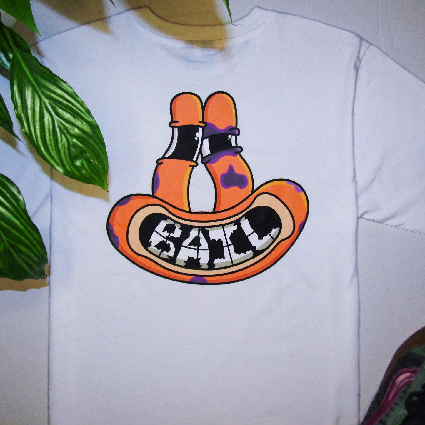 Lifestyle photograph featuring a white graphic T-shirt. Printed on the apparel is a colourful graphic design of an orange frog-like face with tall eyes. The face is smiling, revealing "BAIL" text compiled of shattered teeth, and has several purple bruises, one of which forms a black eye. The apparel is flat-laid on a white background framed by green foliage and a pair of green and purple Vans Knu Skool skating shoes.
